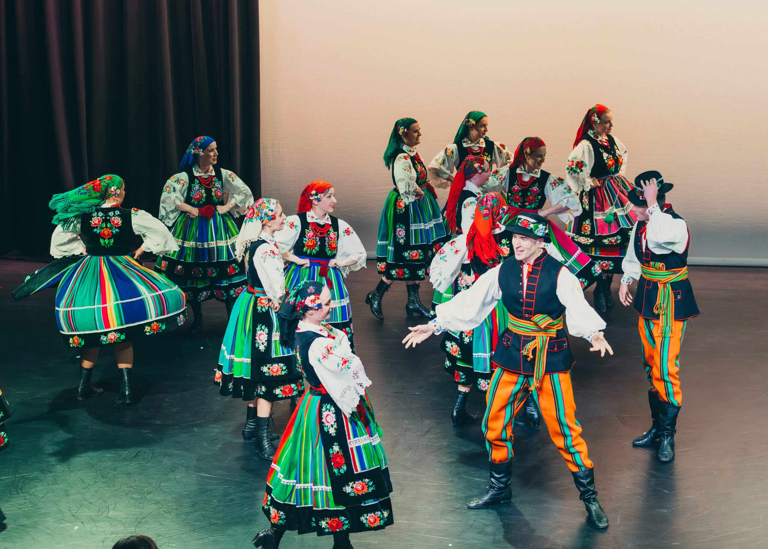 Group of performers in colorful traditional Polish folk costumes dancing on a stage.