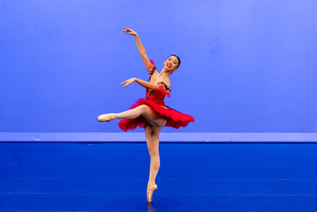 Ballet dancer on pointe in a red tutu performing an arabesque against a blue backdrop