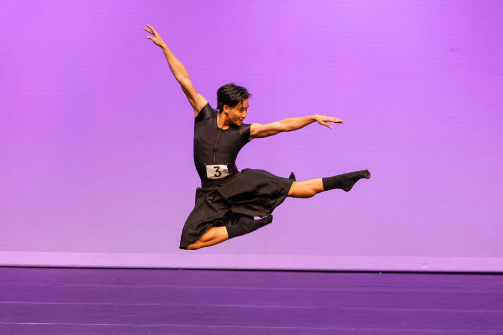 Dancer in black costume performing a mid air leap grand jeté on a purple stage wearing number 3