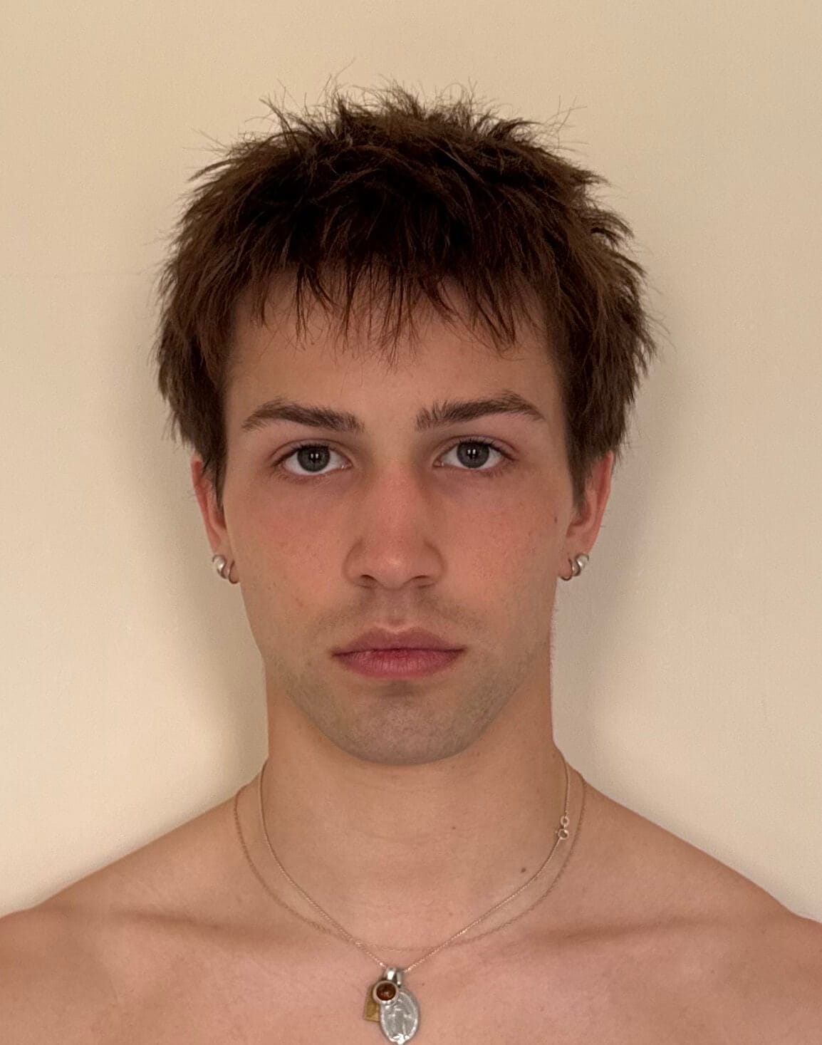 Portrait of a young man with short tousled brown hair and small earrings facing the camera against a beige backdrop wearing layered necklaces
