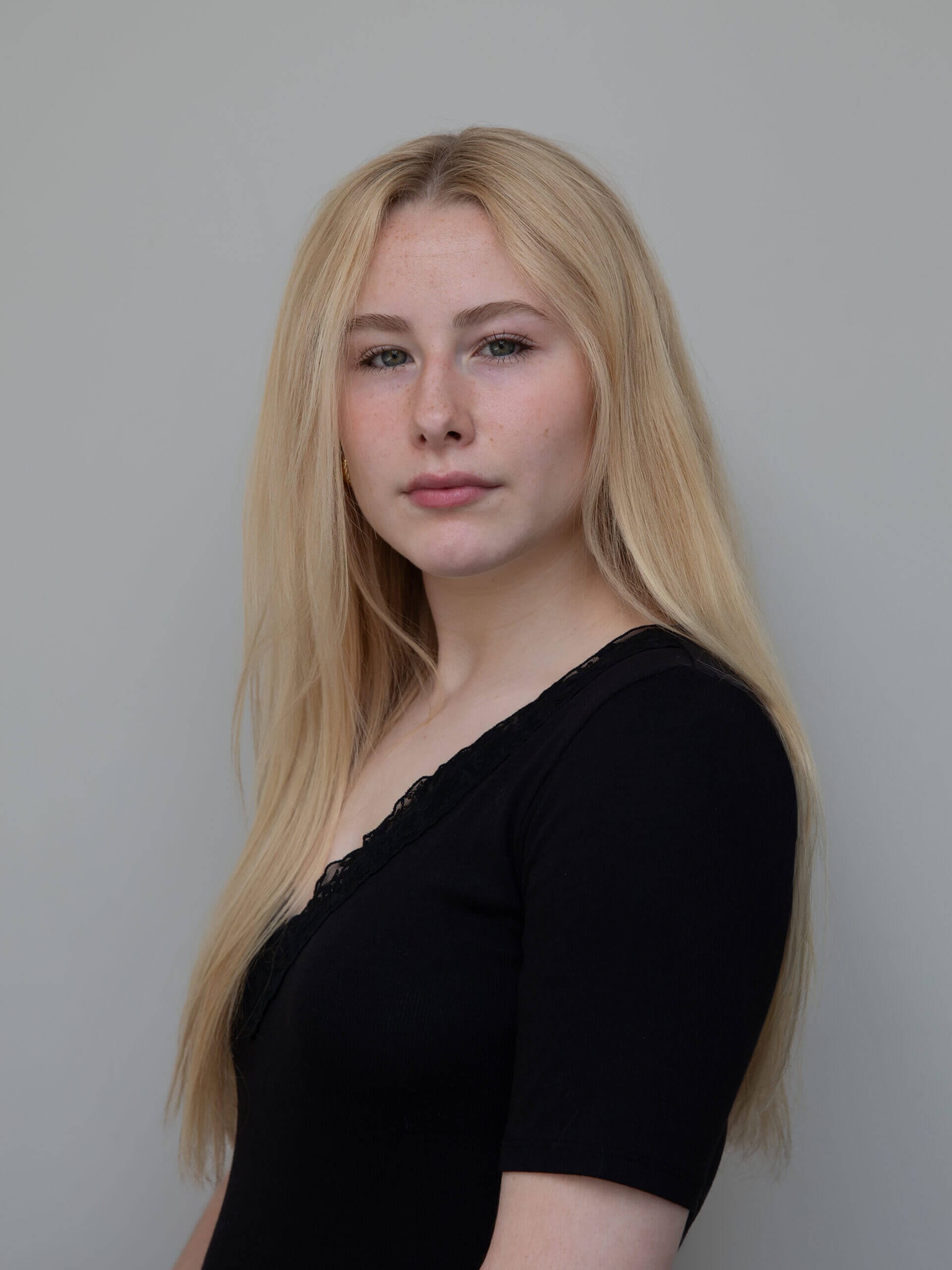 Portrait of a young woman with long blonde hair wearing a black top looking at the camera against a gray background