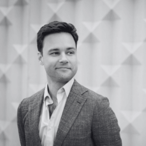 Portrait of a man in a grey blazer and white shirt looking to the right against a geometric wall in a professional setting