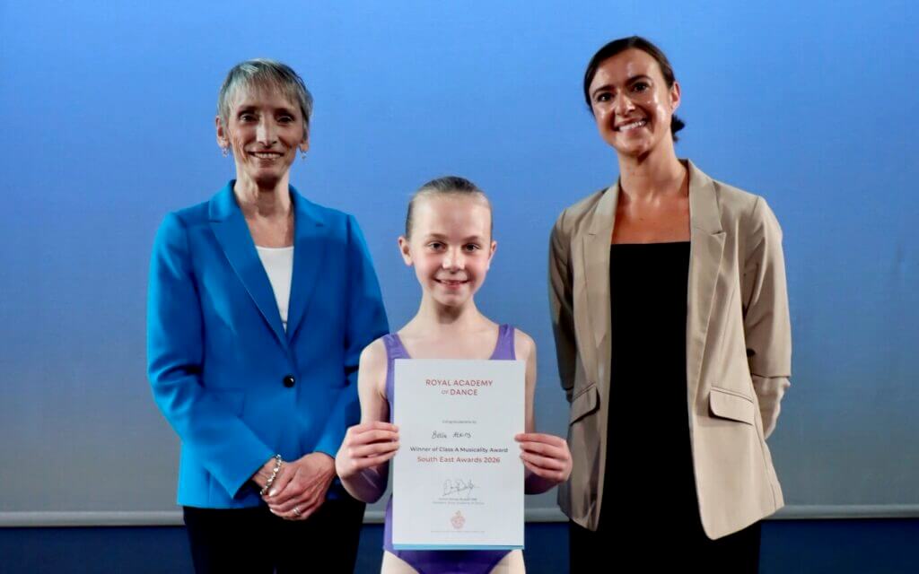 Young girl in a purple ballet outfit holding a certificate flanked by two smiling women on a stage with a blue backdrop