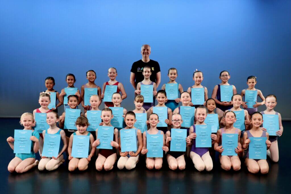Group of young ballet students posing on stage each holding a blue certificate with a smiling man in a black shirt standing behind them against a blue backdrop