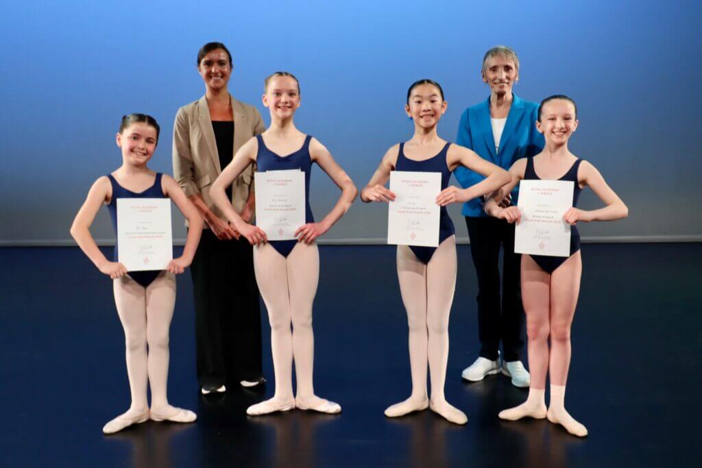 Group of five young ballet dancers on stage each in leotards and tights holding certificates flanked by two instructors in business attire