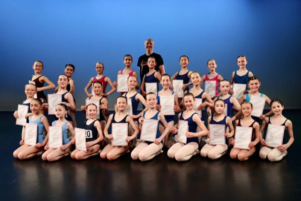 Group of young ballet students posing with certificates on stage instructor standing behind them