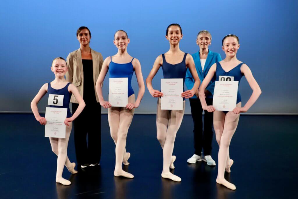 Group of smiling ballet dancers on a stage holding certificates with an adult supervisor in the back