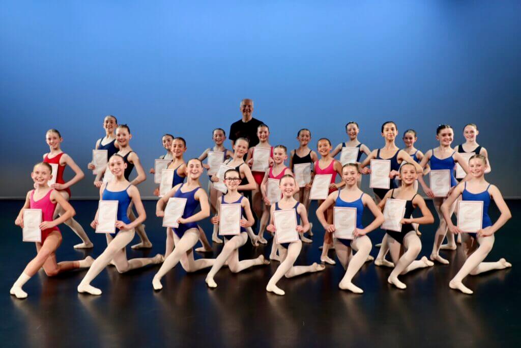 Group of smiling young ballet dancers holding certificates on a stage with a blue backdrop led by an instructor in the center