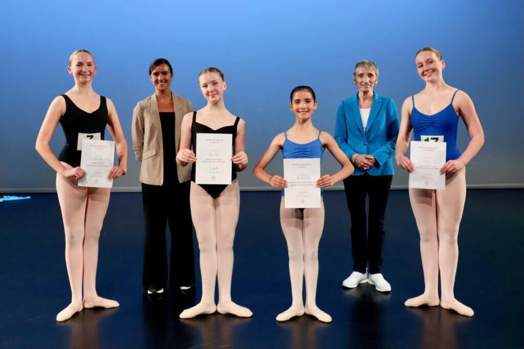 Six ballet dancers stand on a stage each holding a certificate two adults in smart outfits stand behind them against a blue backdrop smiling