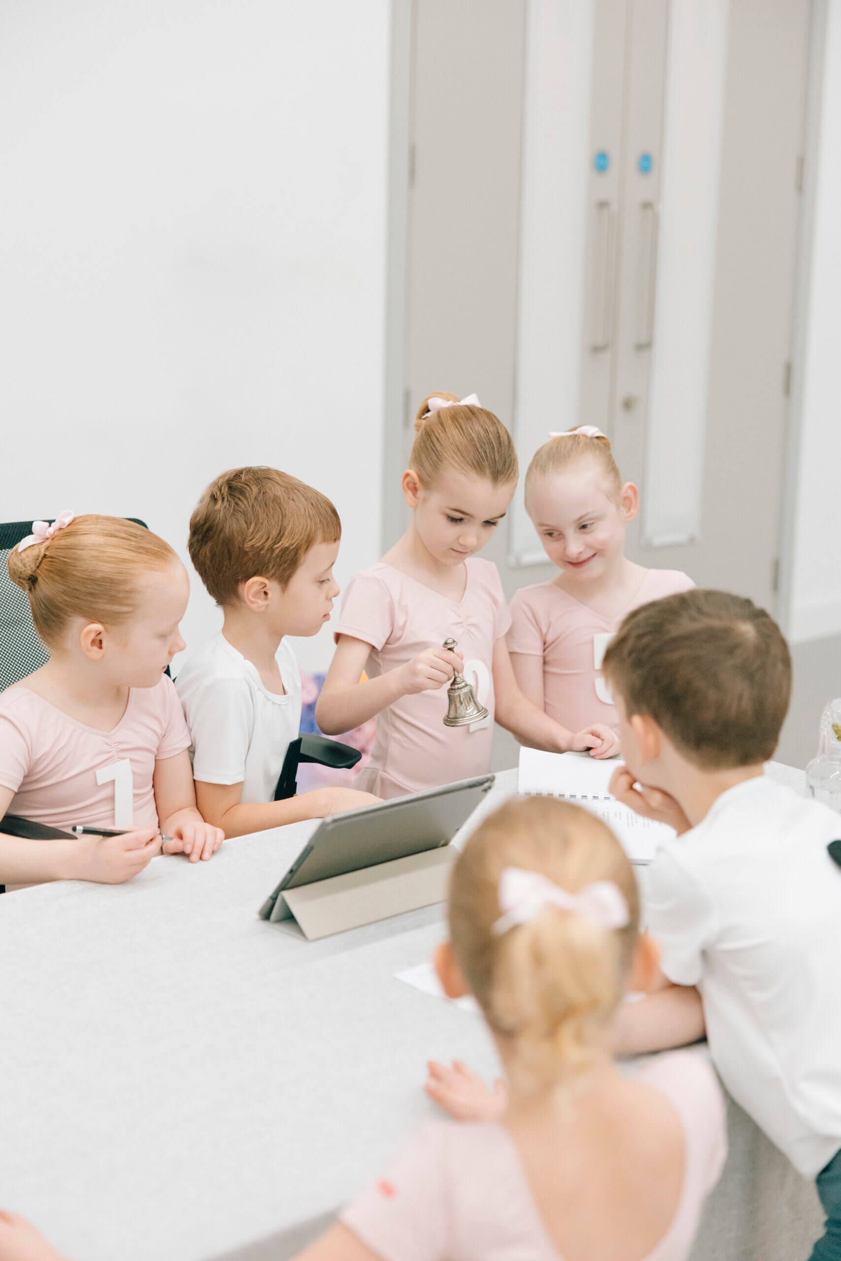 Group of young children in pink outfits gathered around a table in a classroom, with a bell and papers on the desk.