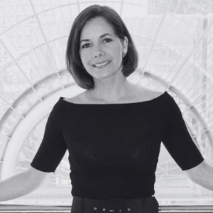 Smiling woman with short dark hair wearing a black boat neck top stands with arms outstretched in front of an ornate arched background