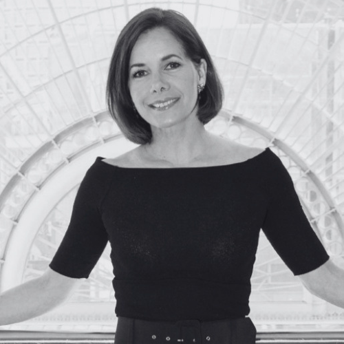 Smiling woman with short dark hair wearing a black boat neck top stands with arms outstretched in front of an ornate arched background