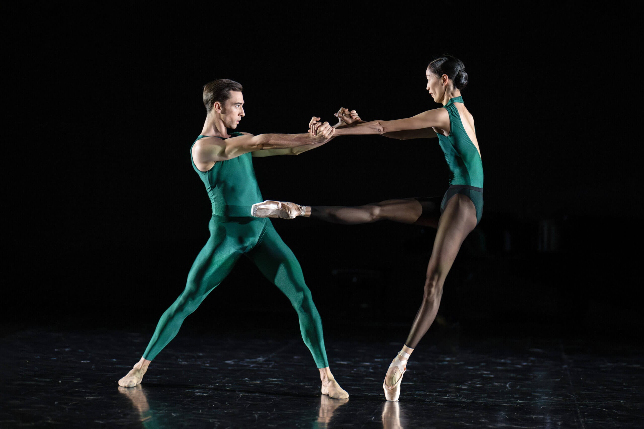 Two ballet dancers in green leotards perform a synchronized partner pose facing each other with arms extended and one leg raised in a lift on a dark stage