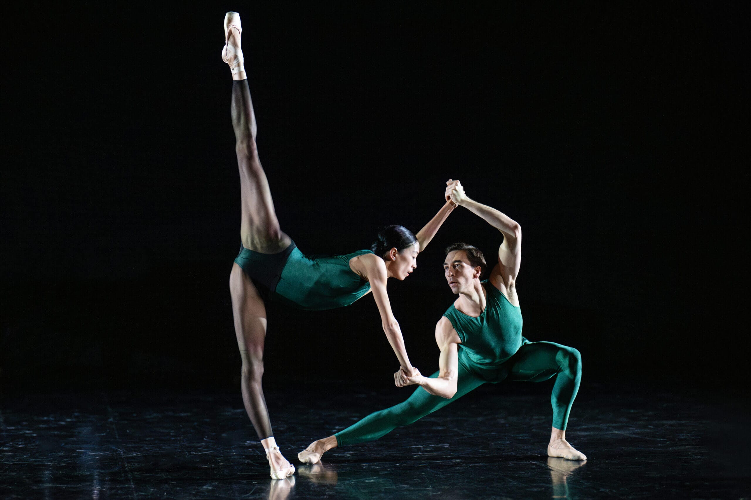 Ballet dancers in green leotards perform a synchronized routine on a dark stage with one dancer's leg raised high behind them.