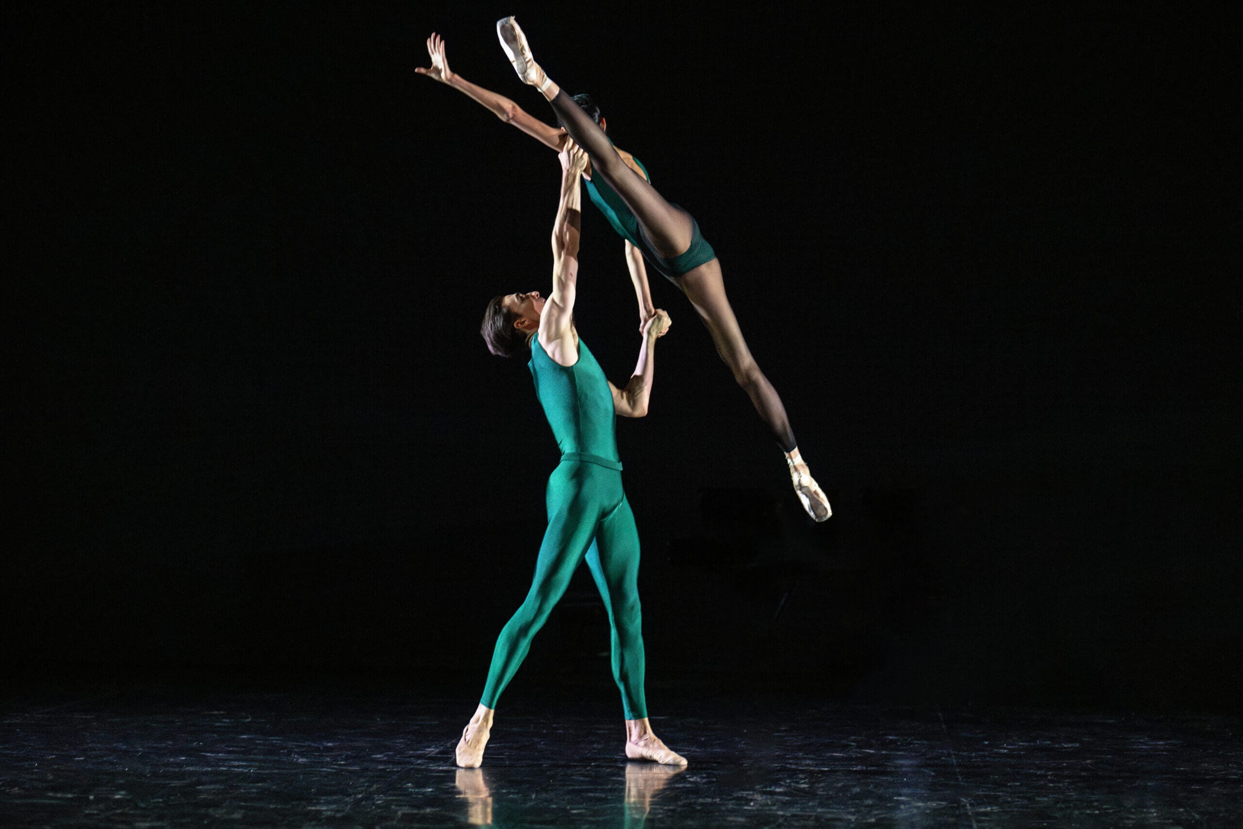 Two dancers in teal perform a dramatic lift on a dark stage one dancer elevated horizontally above the other