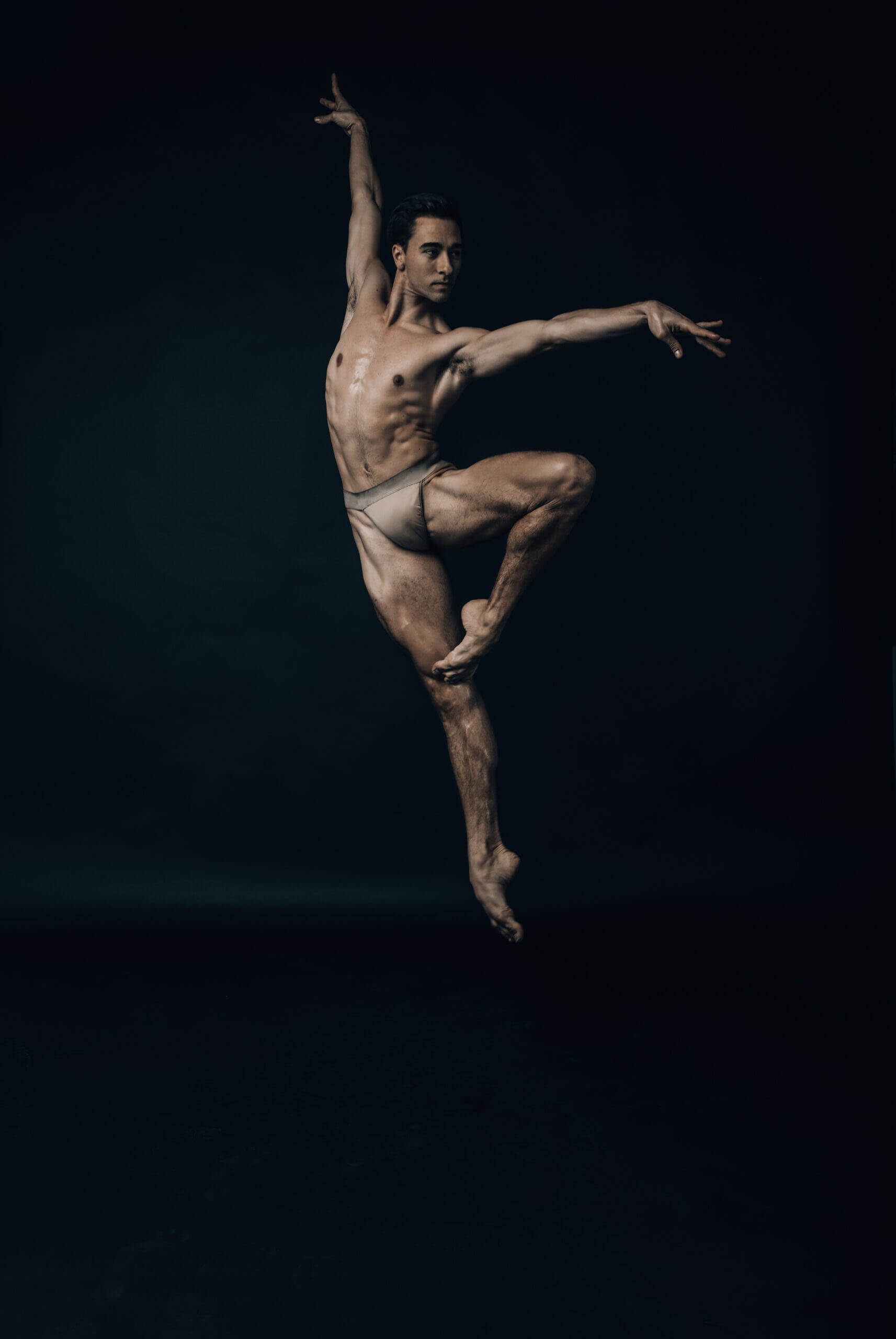Male ballet dancer leaping in a grand jeté pose arms extended wearing light tights against a dark background