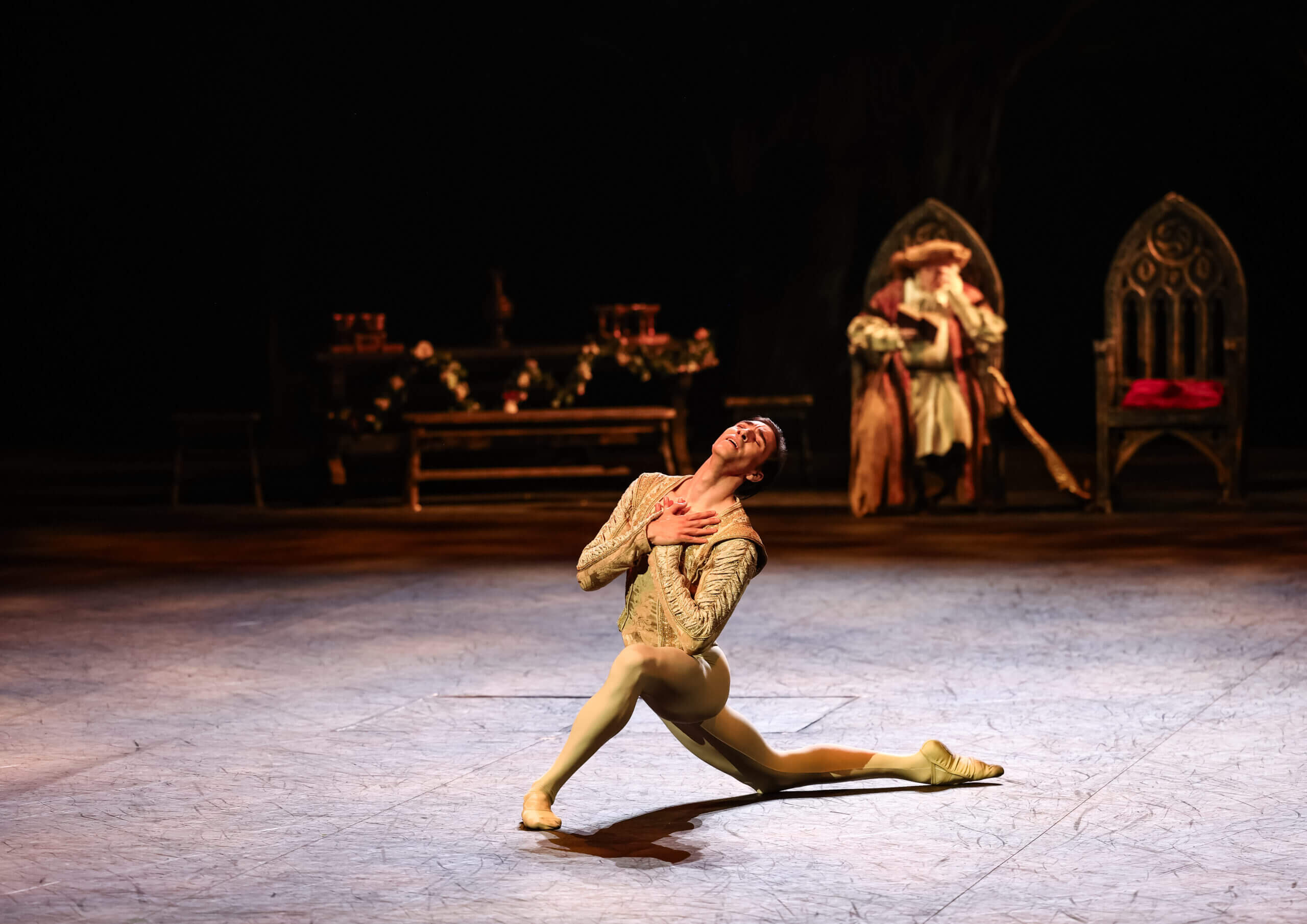 Dancer in a gold costume performs a dramatic split on stage with ornate throne props and floral decor in the background