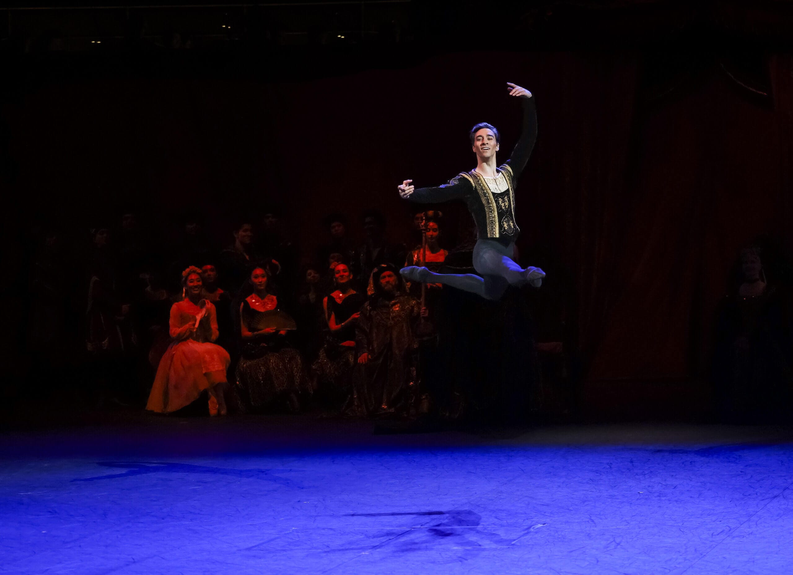 Male ballet dancer in a gold embroidered costume leaps mid air on a dark stage with a blue lit floor and an audience in the background