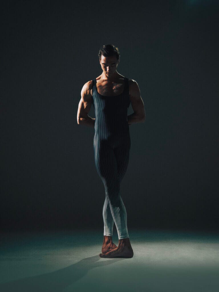Person wearing a dark pinstriped bodysuit stands with hands behind their back on a dim stage