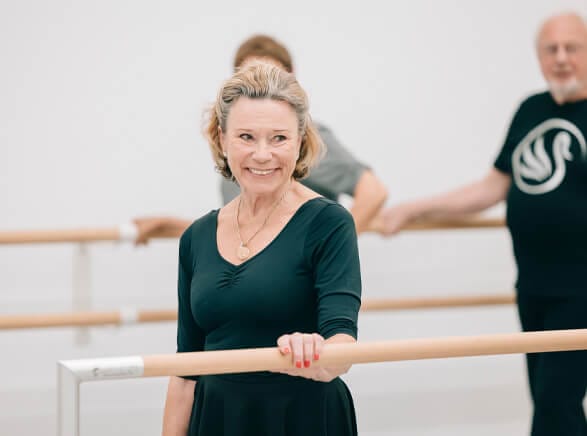 Smiling older woman at a ballet barre in a dance studio practicing with others in background