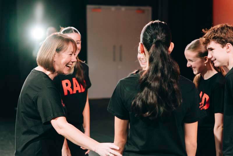 Instructor shakes hands with a student in a black tee while four others smile in a backstage rehearsal space stage lights on