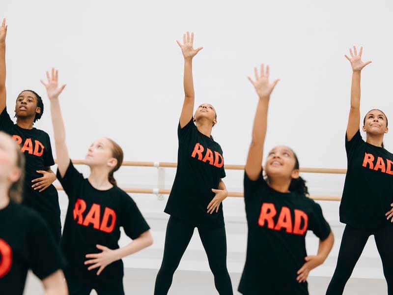 Group of kids in a dance studio raising their arms upward in a choreographed pose wearing black shirts that read RAD