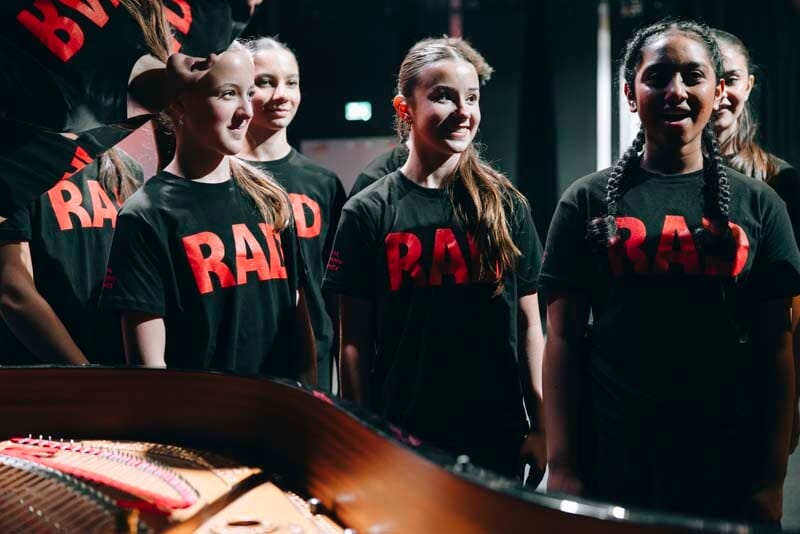 Group of young performers in black T shirts with large red lettering standing near a grand piano backstage