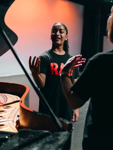 Young woman in a black shirt with red lettering speaks and gestures toward an open grand piano in a music studio