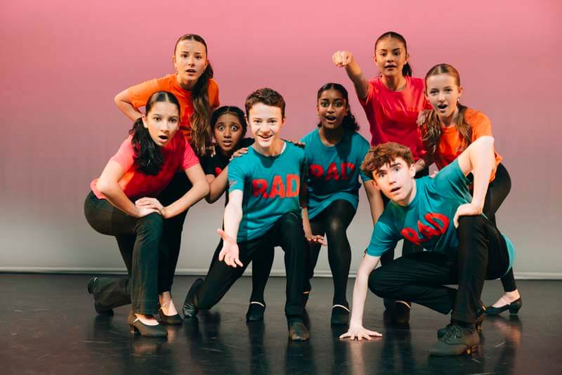 Group of diverse kids on stage dressed in teal and bright shirts mid dance pose facing the audience with outstretched arms and focused expressions