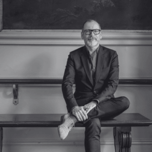 Black and white portrait of a middle aged man in a suit and glasses sitting cross legged on a bench with hands folded