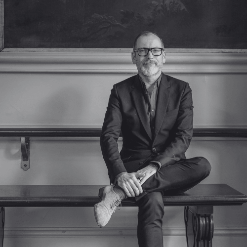 Black and white portrait of a middle aged man in a suit and glasses sitting cross legged on a bench with hands folded