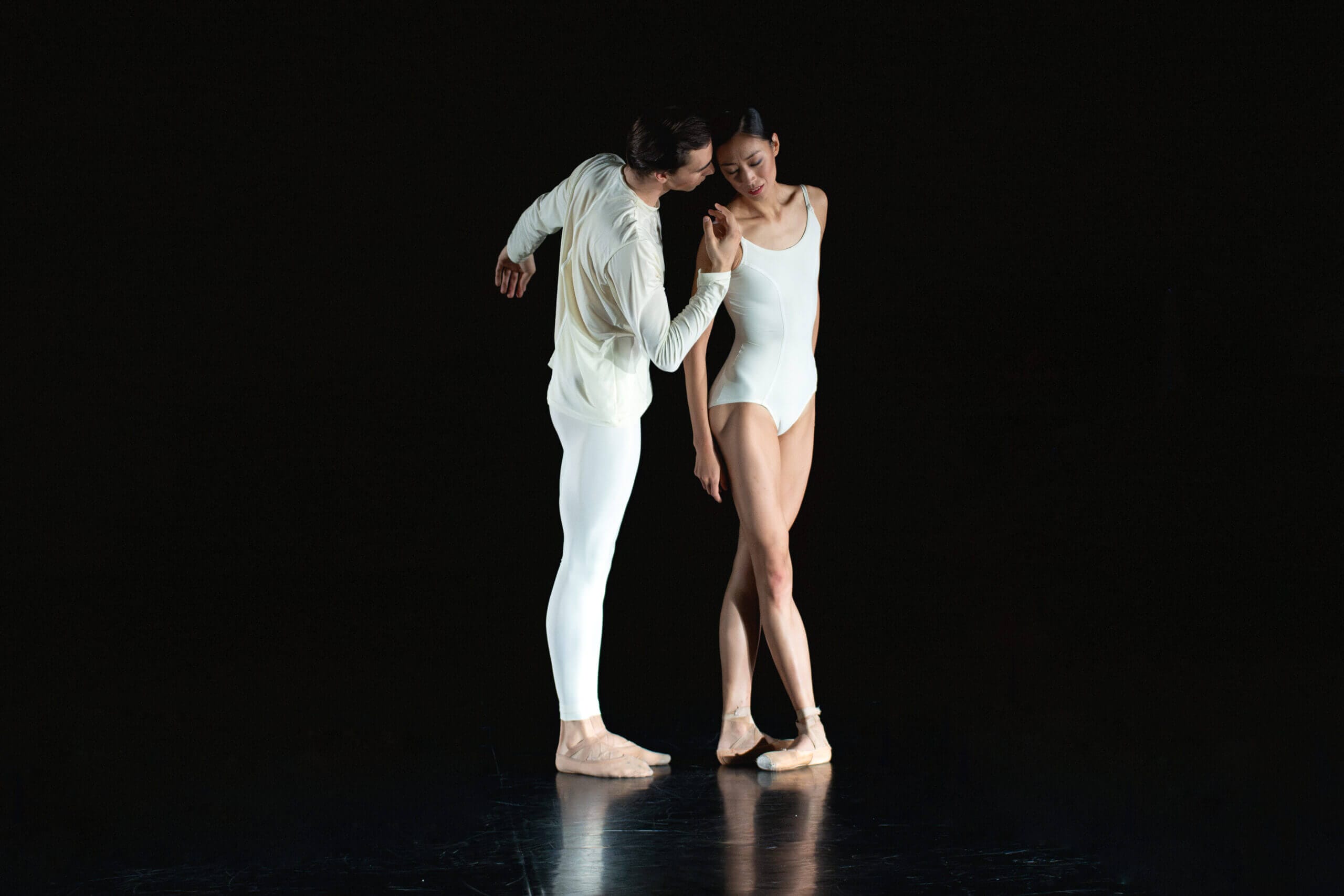 Two ballet dancers in white on a dark stage share a close intimate duet heads near each other and gentle contact at the handsor wrists