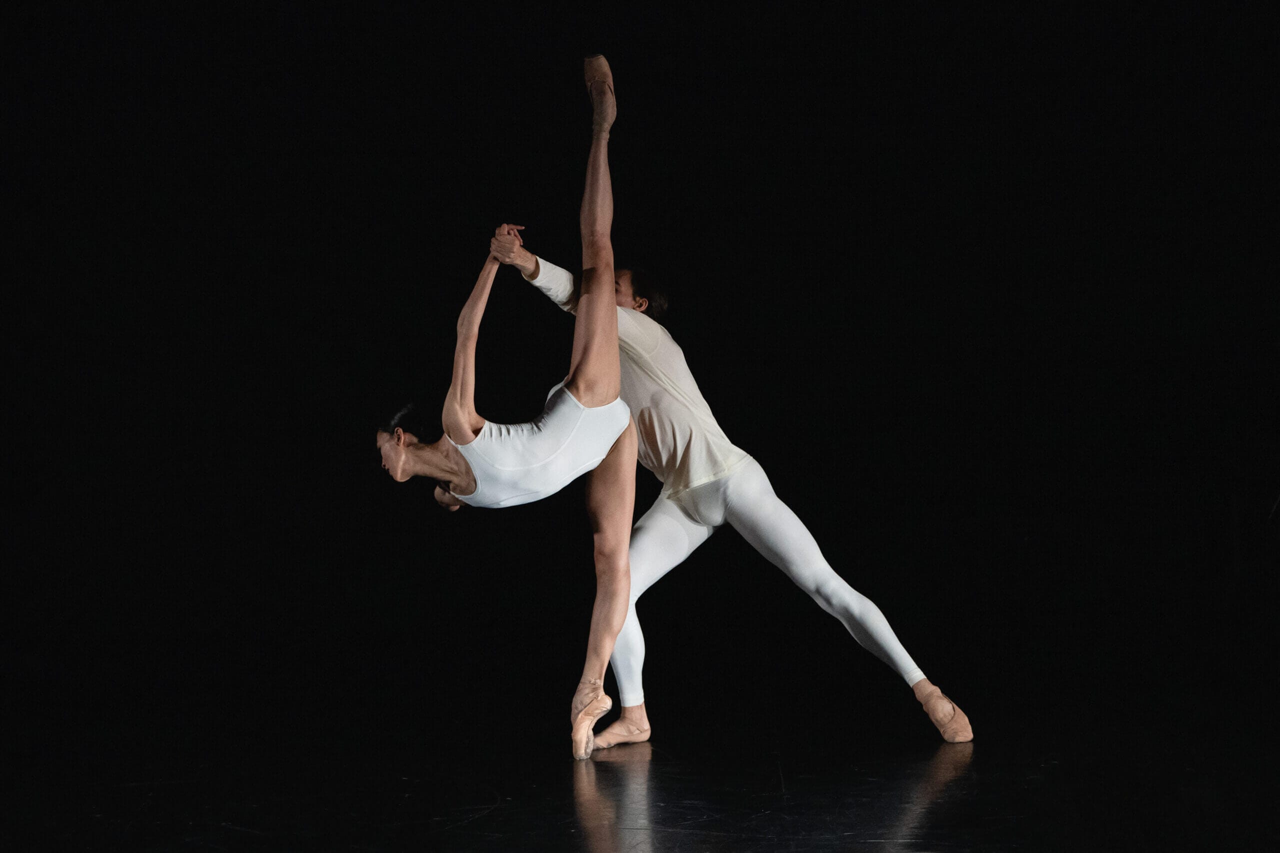 Three dancers in white perform an acrobatic pose on a dark stage one leg raised high as others support and balance the group