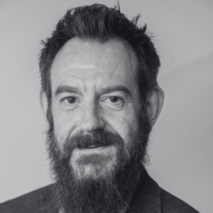 Black and white portrait of a middle aged man with a full beard and short hair looking at the camera