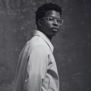 Black and white portrait of a person with short curly hair and glasses wearing a light blazer and looking over their shoulder at the camera