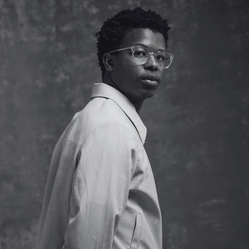 Black and white portrait of a person with short curly hair and glasses wearing a light blazer and looking over their shoulder at the camera