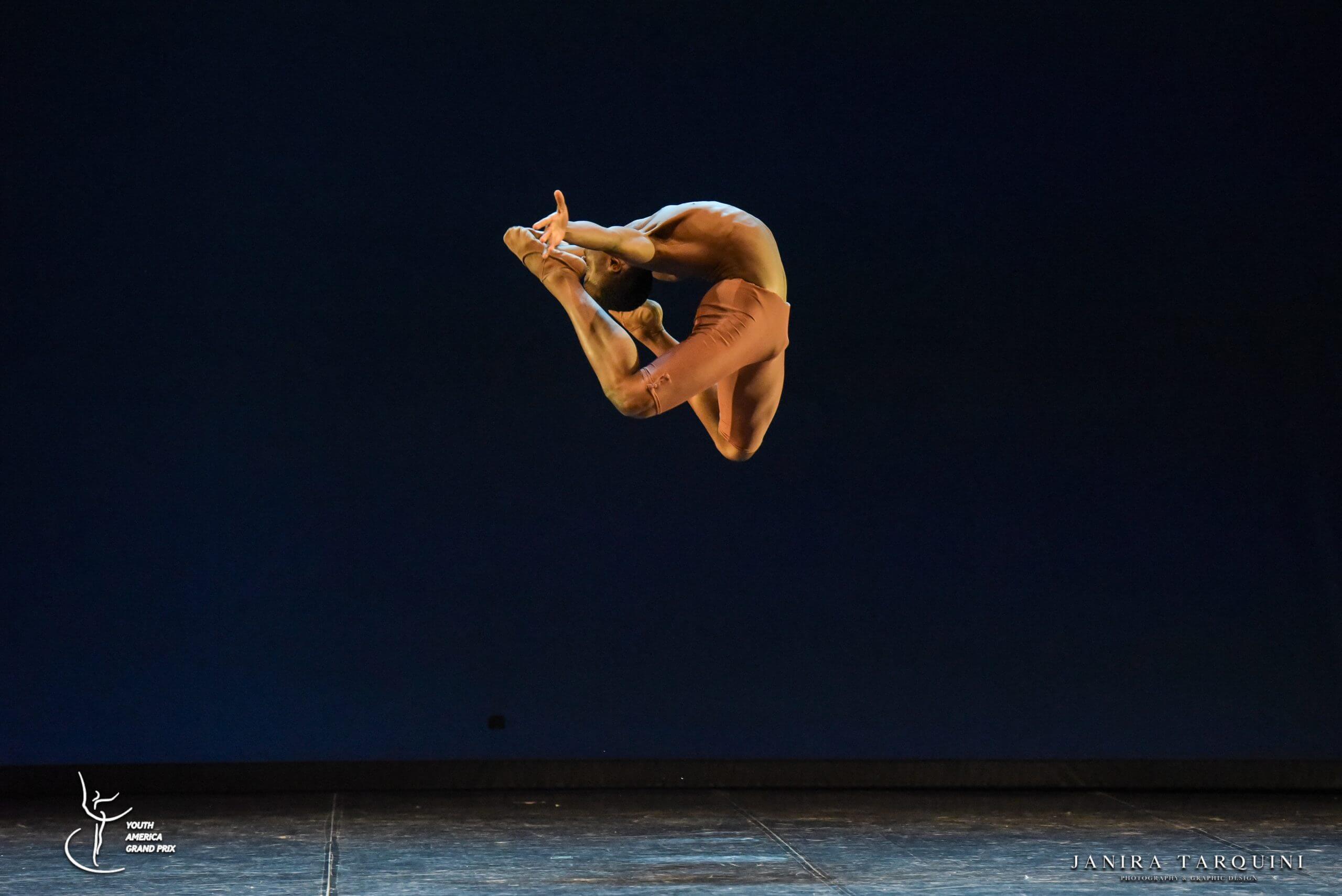 Dancer in orange tights leaps mid air on a dark stage with stage floor visible at the bottom and logos in the corners
