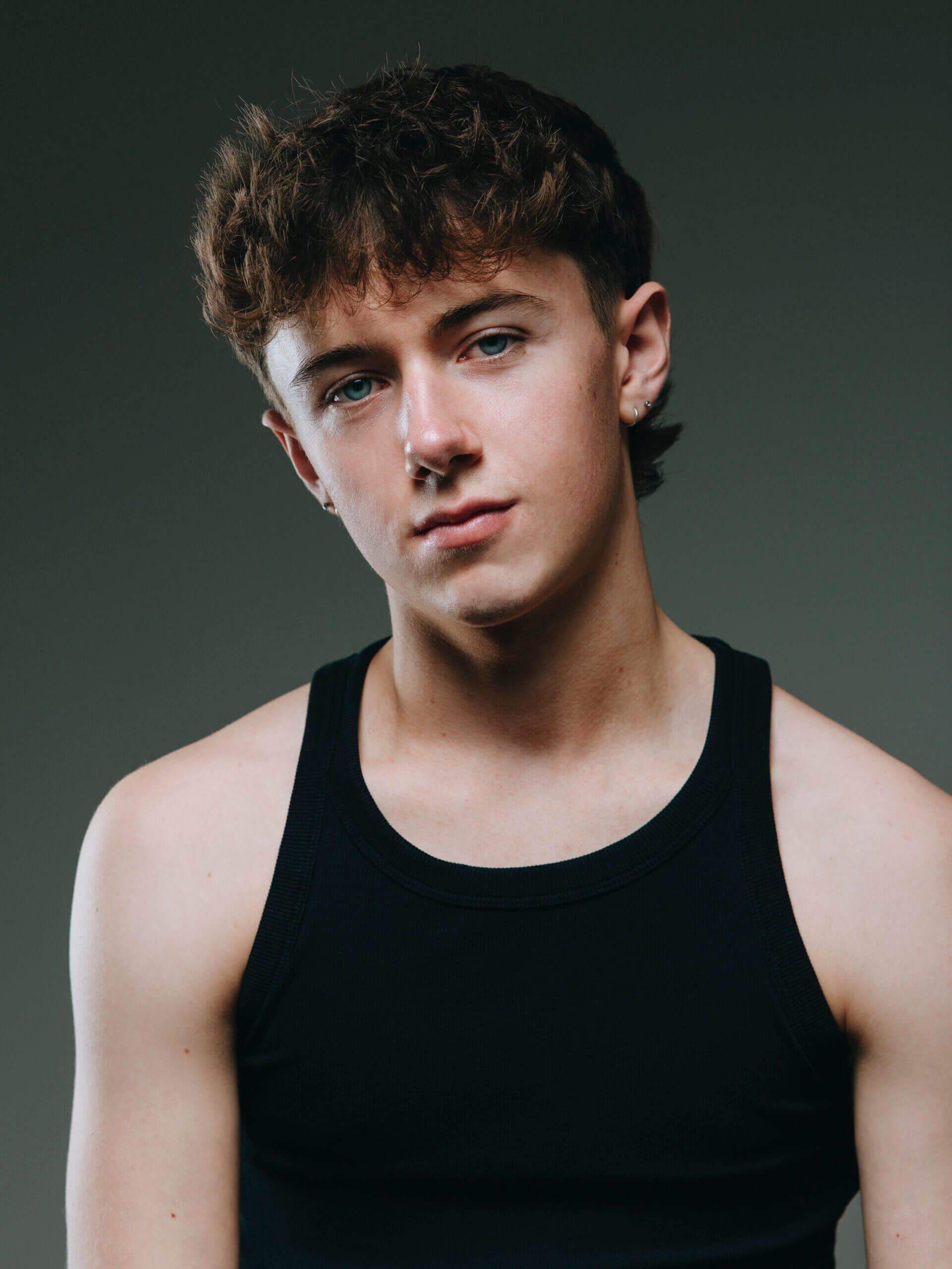 Close up portrait of a young man with blue eyes wearing a black tank top head slightly tilted against a dark gray background