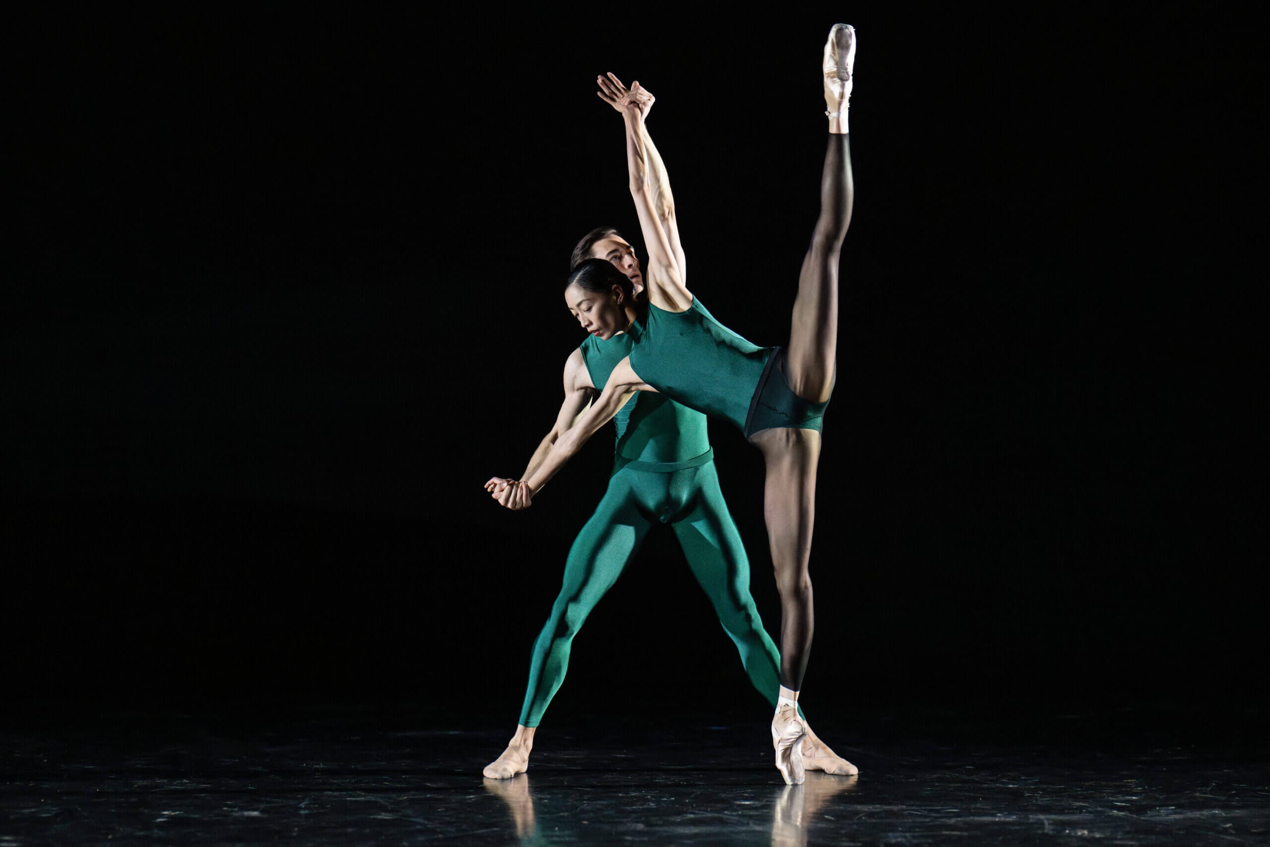Two dancers in teal perform a high leg extension on a dark stage