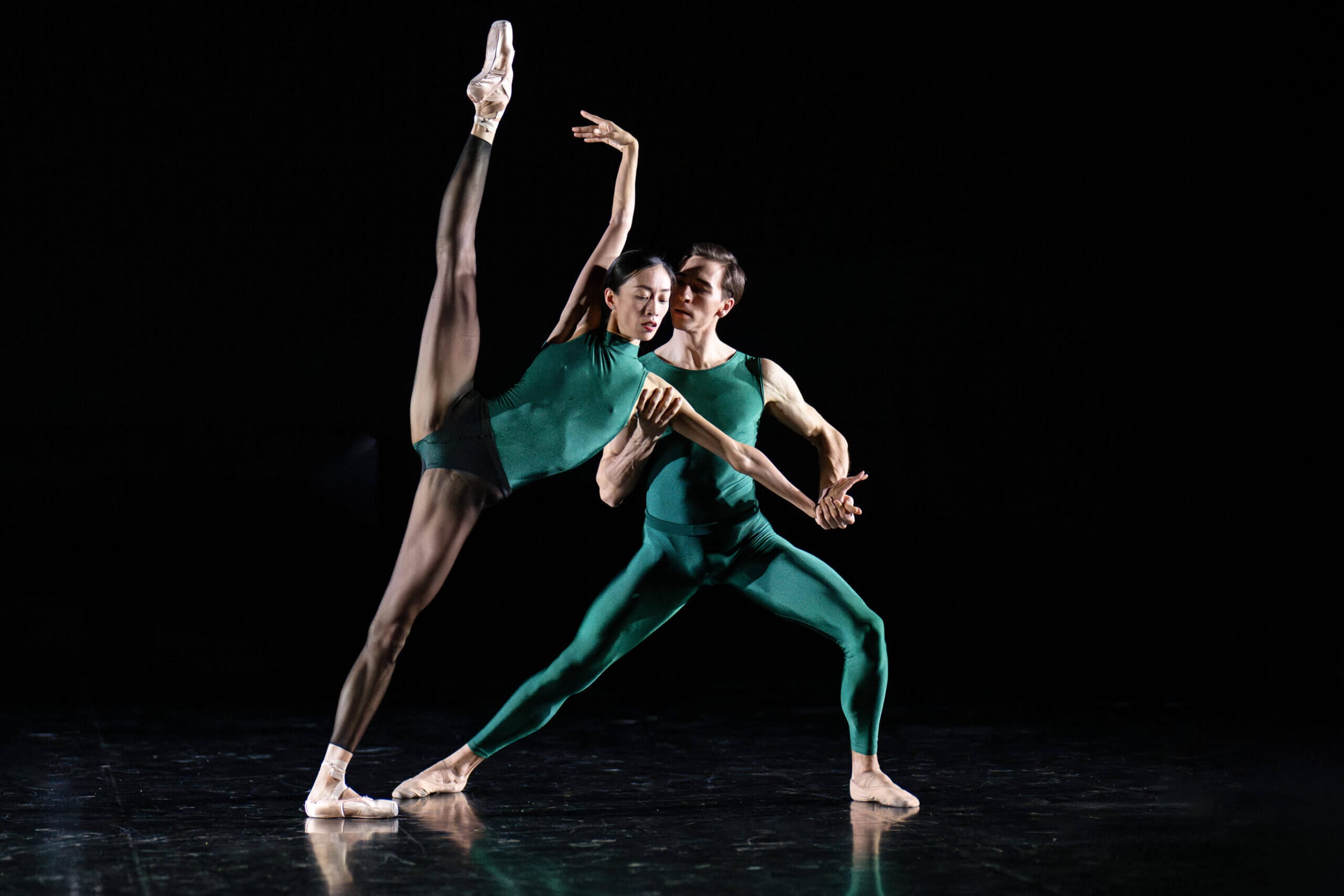 Two ballet dancers in teal green outfits perform a duet on a dark stage one dancer raises a leg into a high arabesque while the other supports the pose