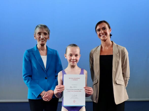 Young girl in a purple dance leotard holds a certificate as two smiling women in blazers stand beside her on a blue stage backdrop