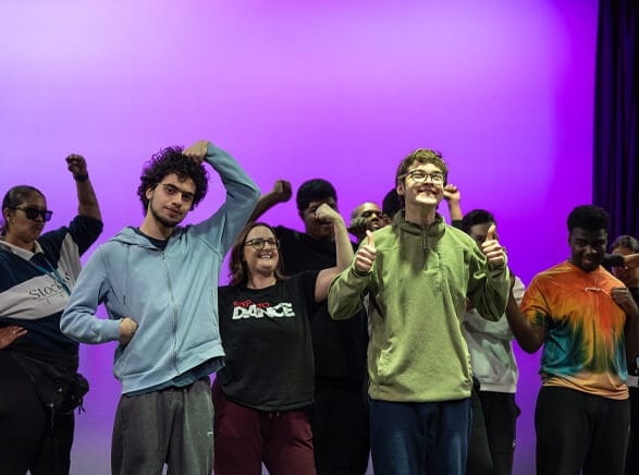 Group of diverse teens on a stage with a purple backdrop giving thumbs up and smiling