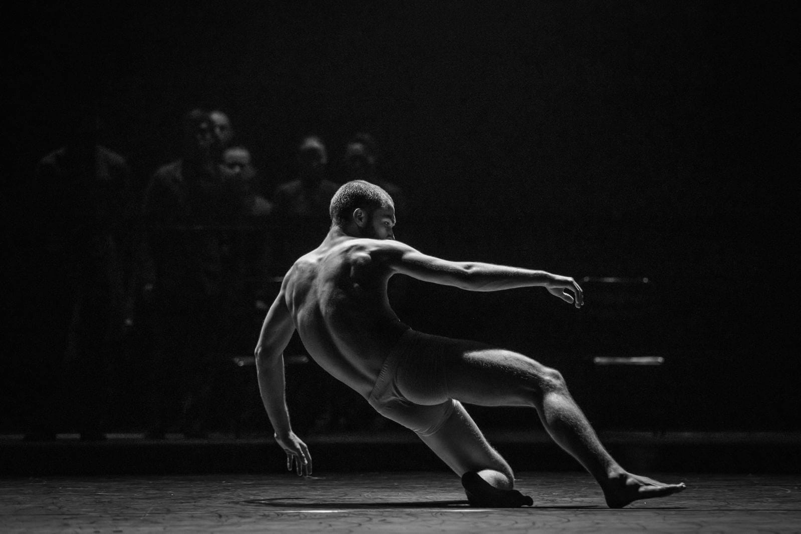 Male dancer on a dimly lit stage shirtless reaching his right arm forward in a dramatic pose with audience in the background black and white