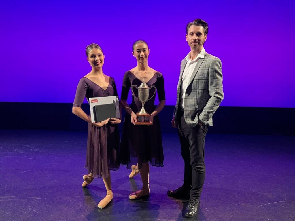 Three dancers stand on a stage with a purple background two women in brown dance gowns hold a trophy and a certificate while a man in a gray plaid blazer stands beside them posing for a photo